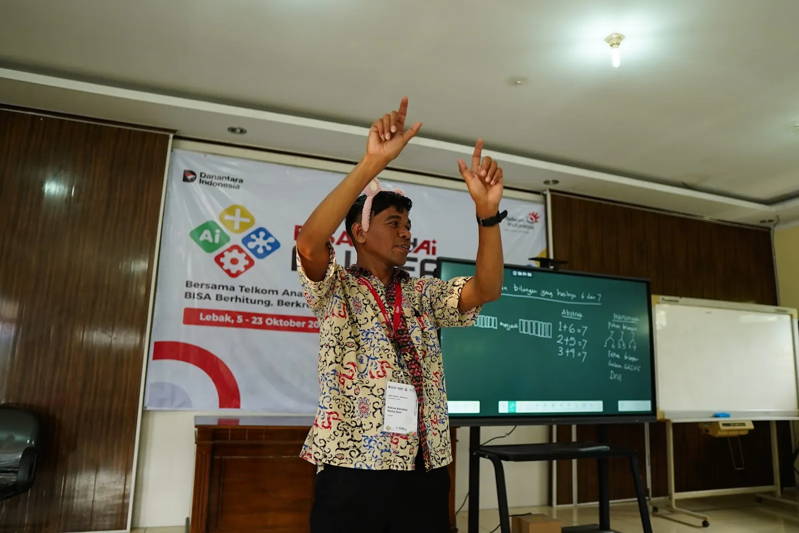 Fasilitator mengangkat kedua tangan di depan papan bertuliskan penjumlahan yang hasilnya tujuh