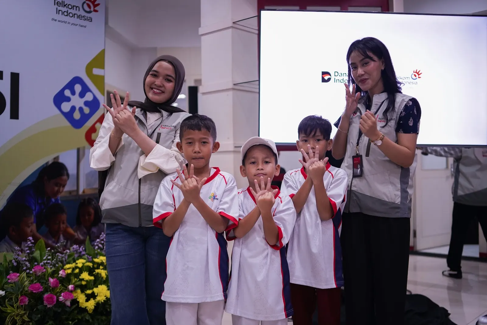 Tiga siswa dan dua fasilitator memperagakan isyarat jari yang sama di depan panggung